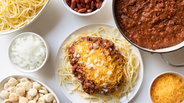 A plate of spaghetti topped with chili and cheese, surrounded by ingredients in individual bowls
