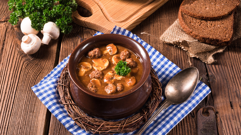 A bowl of mock turtle soup served on a blue and white checkered napkin with sliced bread, whole mushrooms, and fresh herbs
