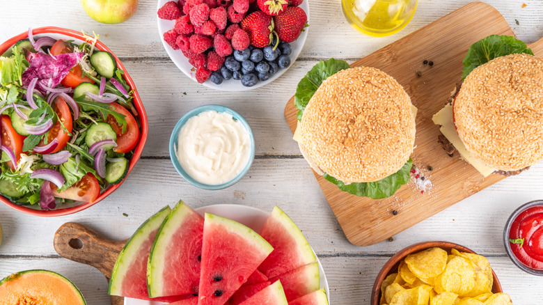 Burgers, salads, chips, and other typical cookout foods spread out over a wooden table.