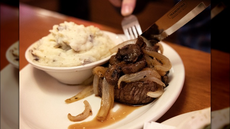 Cutting steak with grilled mushrooms and onions plus a small bowl of mashed potatoes on the side.