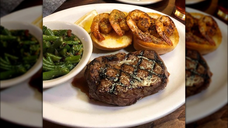Juicy steak on plate with dinner rolls, shrimp, and small bowl of green beans.