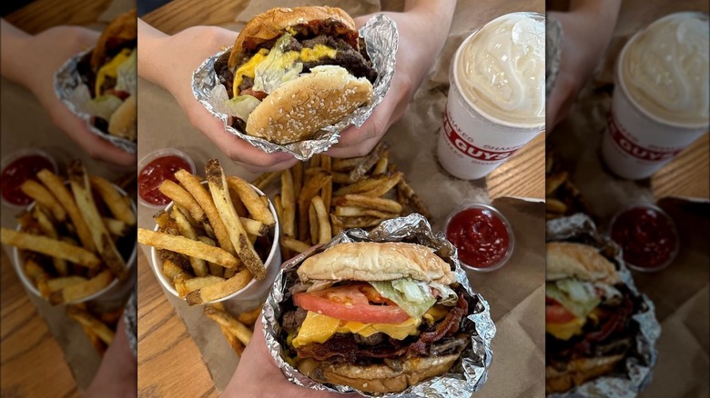 Two people grip foil wrapped burgers surrounded by beverages, fries, and ketchup.