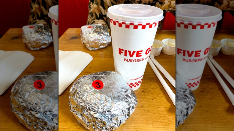 Two wrapped sandwiches and a beverage on a table at a Five Guys restaurant.
