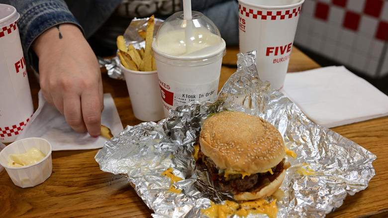 Person at Five Guys restaurant with cheeseburger, fries, milkshake, and beverages.