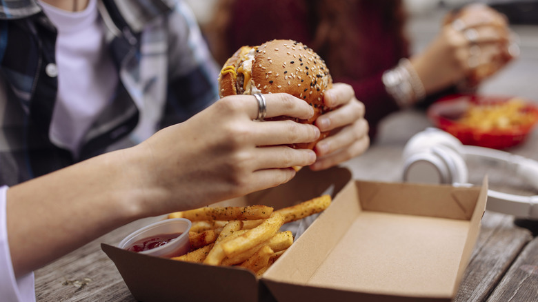 People eating fast food burgers and fries on wooden table outdoors.