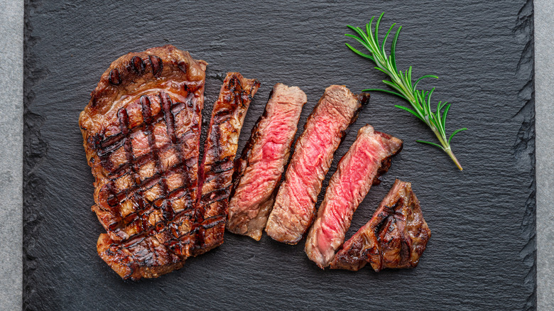 A grilled, sliced, medium-rare steak on a slate cutting board, a sprig of rosemary to the side.