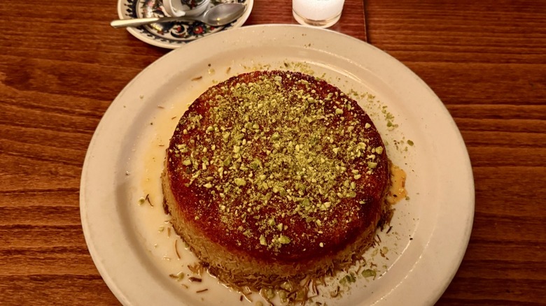 Tanoreen's round knafeh dessert topped with green pistachios on a white plate