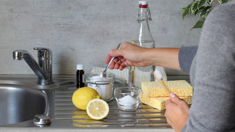 Person holding spoon in glass jar with white powder on counter next to sink with lemons and sponges.