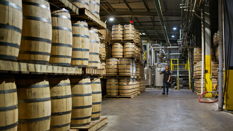 A worker inspecting stacked rows of whiskey barrels in a warehouse.
