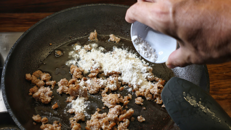 hand dumping small dish of flour into skillet of crumbled sausage