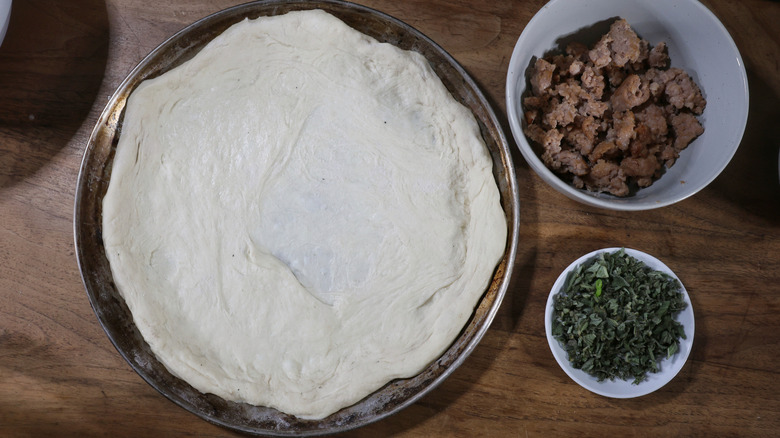 Pizza dough spread on small pan with bowls of toppings beside it