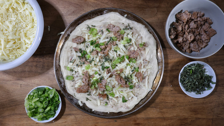 uncooked sausage and gravy breakfast pizza on table with bowls of toppings beside it
