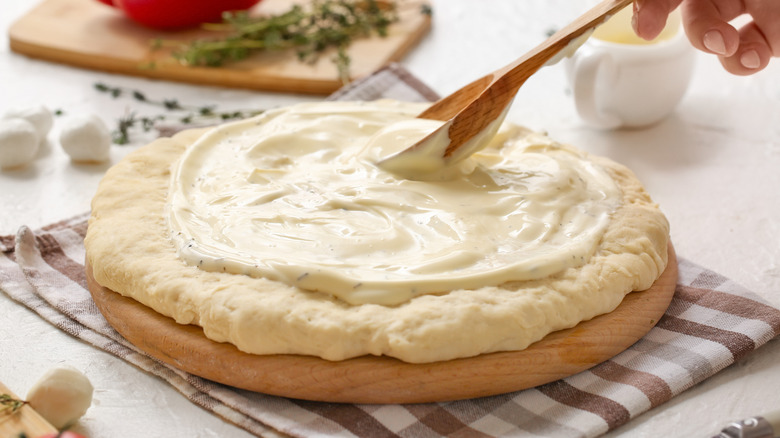 Hand uses spoon to spread white sauce on pizza dough
