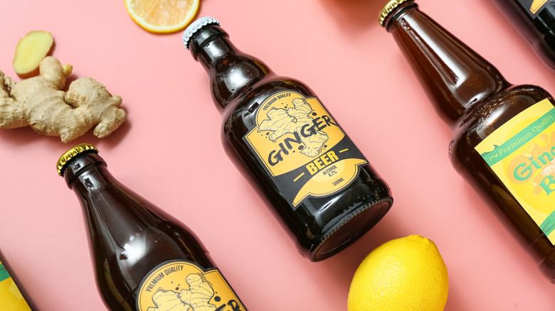 Three bottles of ginger beer on pink background with ginger root and lemons