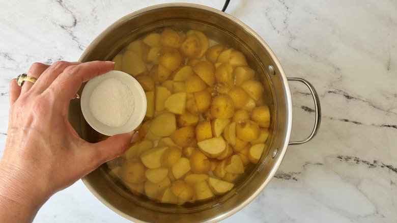 Hand holding small bowl of salt over potatoes boiling in pot