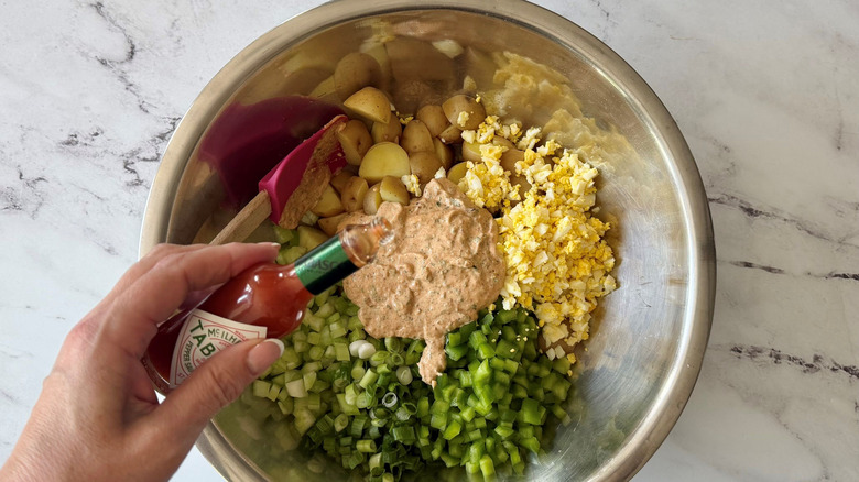 Hand holding Tabasco bottle over unmixed potato salad in bowl