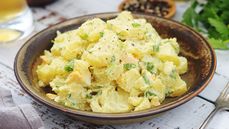 Potato salad in bowl on wooden table with fork, napkin, and ingredients in background