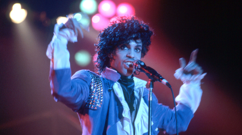 Prince singing in front of a microphone with his arms up in the air and a backlit stage behind him