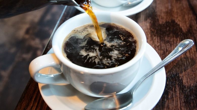 Coffee being poured into a white mug, atop a saucer and next to a spoon
