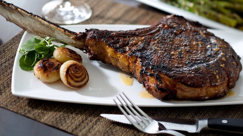The Capital Grille bone-in tomahawk ribeye steak on a plate with rolls and cutlery
