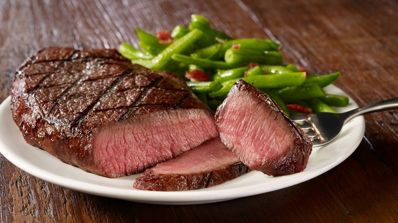 fork holds a bite of steak in front of a plate of steak and green beans