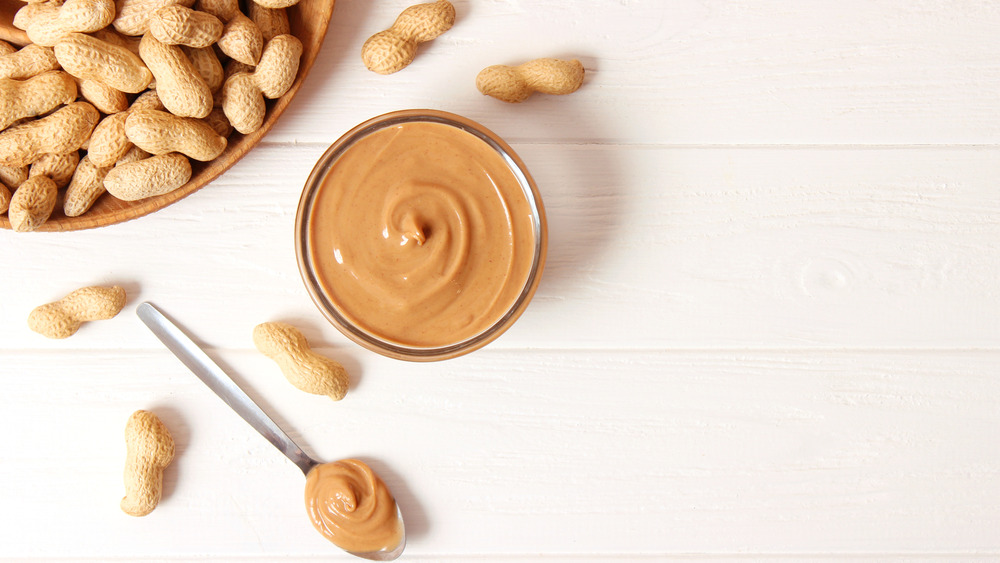 peanut butter in a jar and on a spoon near a bowl of shelled peanuts on a white table