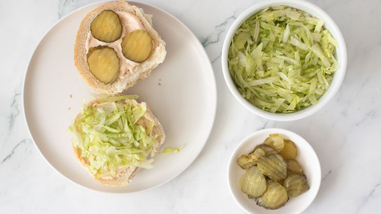 Buns with pickles and lettuce next to bowls of pickles and lettuce