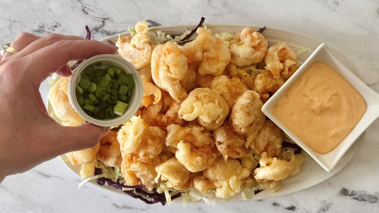 Hand placing small bowl of scallions onto platter of tempura shrimp