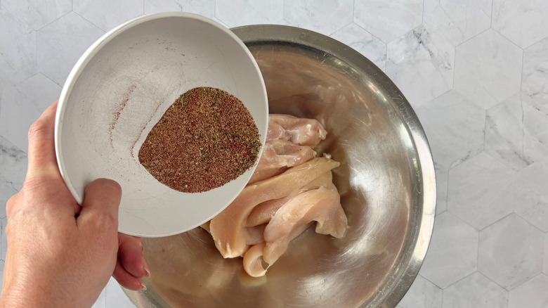 hand holding bowl of seasoning mix over bowl of raw chicken