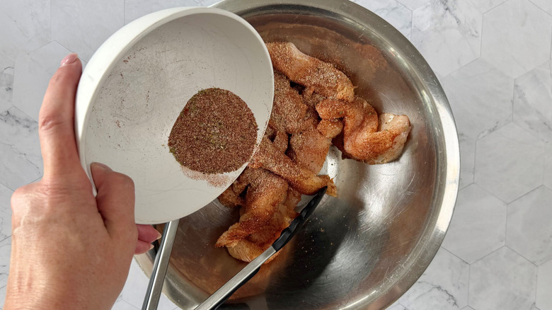 hand holding bowl of seasoning mix over bowl of raw chicken with tongs