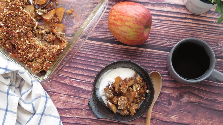 bowl of apple crisp with cup of coffee, apple on table next to baking dish of apple crisp