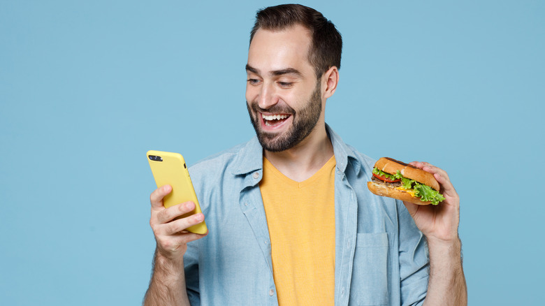 man holding smartphone and hamburger and smiling