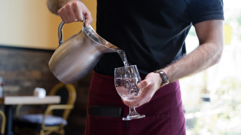 A waiter in a black t-shirt and maroon apron pours water from a metal pitcher into a glass in a restaurant