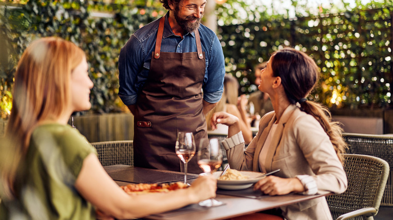 Waiter in denim shirt and brown apron smiles at two customers eating in outdoor restaurant patio