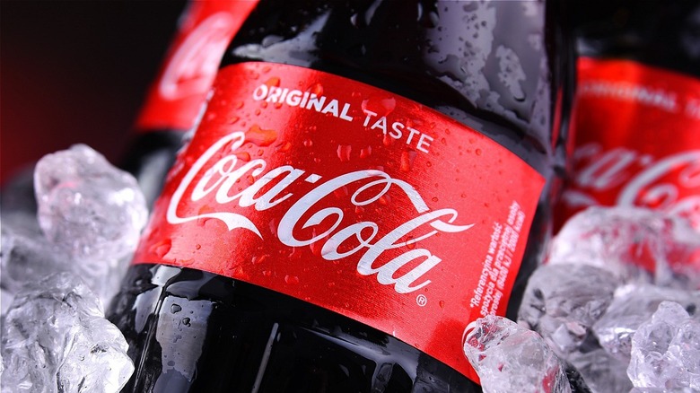 Close up of a bottle of Coca-cola including the label and part of the bottle with other bottles and ice in the background
