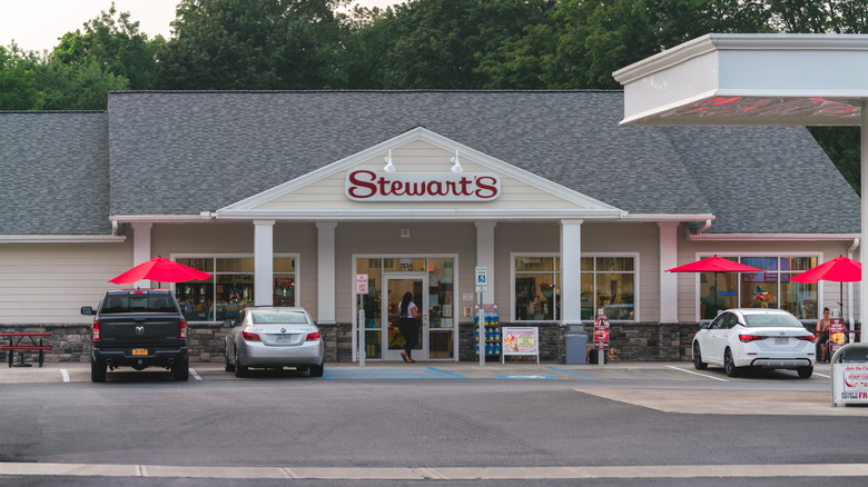 The exterior of a beige Stewart's Shops building with red umbrellas, a gray roof, and cars parked outside