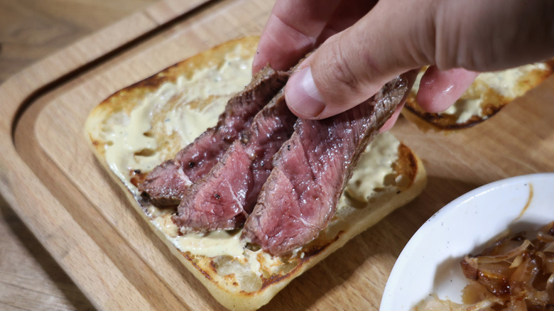 hand placing strips of steak onto a ciabatta roll slathered in Dijonnaise