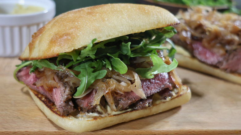 steak sandwich on ciabatta roll with caramelized shallots and baby arugula sitting on cutting board