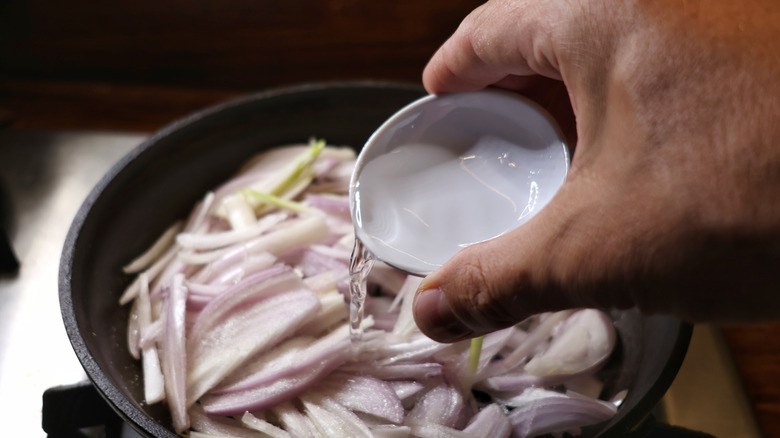 hand pouring small dish of water into skillet of sliced shallots