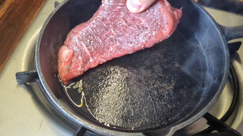 hand placing raw sirloin steak into glistening cast iron pan