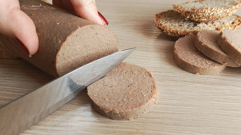 Closeup of hands cutting liverwurst with a metal knife on a wooden surface.