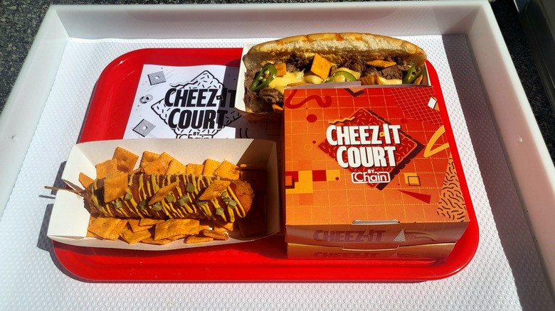 A red tray with three dishes on it: a corn dog sprinkled with Cheez-Its, a cheesesteak sandwich, and a paper box with the words 