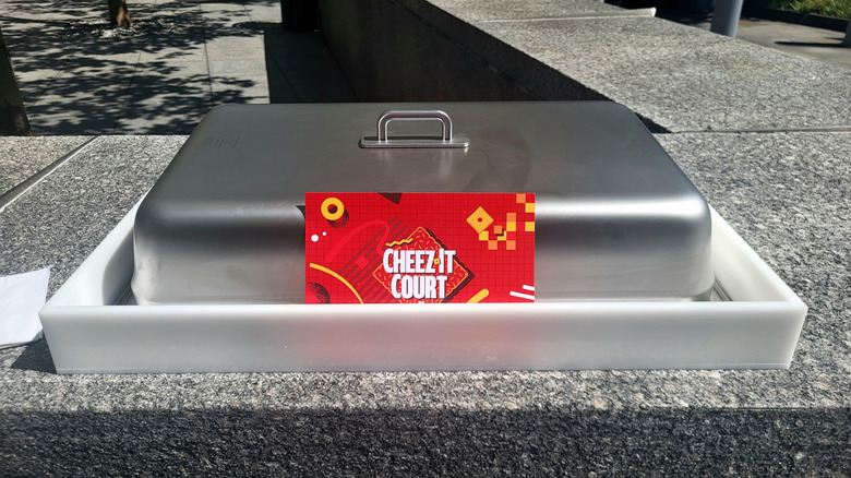 A white tray with a red menu card inside of it reading "Cheez-It Court." The card leans against a metal lid with a handle.