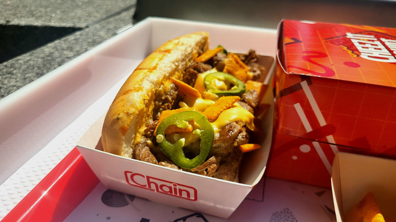 A side view of a cheesesteak sandwich in a paper tray with the word "Chain" printed on it. The sandwich includes sliced beef, melted yellow cheese, rings of jalapenos, and Cheez-Its.