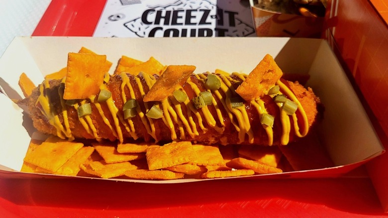 A close-up on the corn dog in a paper tray. It's drizzled with mustard and ketchup, and sprinkled with chopped pickles and Cheez-Its; it's also served on a bed of the crackers.