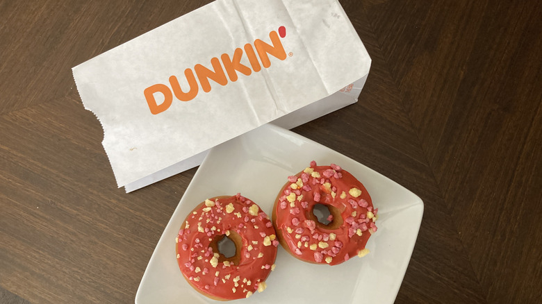 Two Dunkin' fruit punch donuts on a white plate next to the Dunkin' paper bag