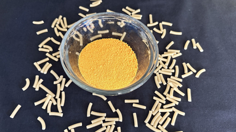 glass bowl of cheese powder surrounded by uncooked noodles on black background