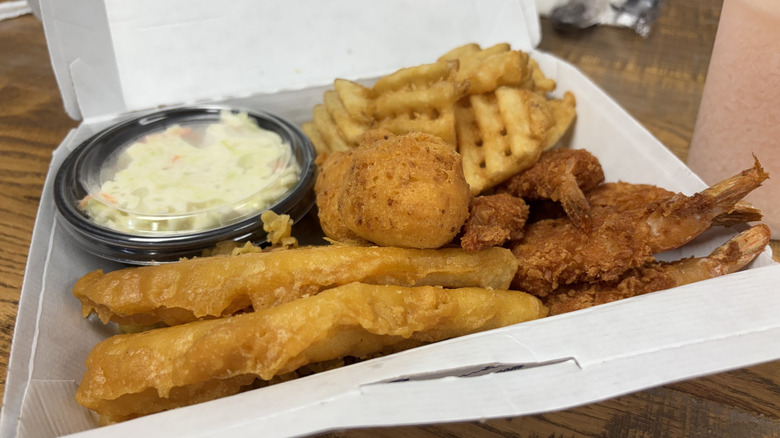 A box of Long John Silver's chicken planks, shrimp, hushpuppies, waffle fries, and cold slaw in container on wooden table