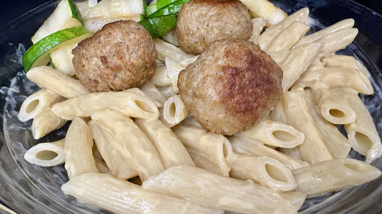 Penne pasta with white sauce, meatballs, and zucchini in a glass bowl
