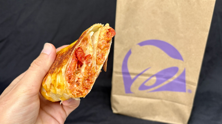 Hand holding half a grilled cheese burrito with Taco Bell bag in background